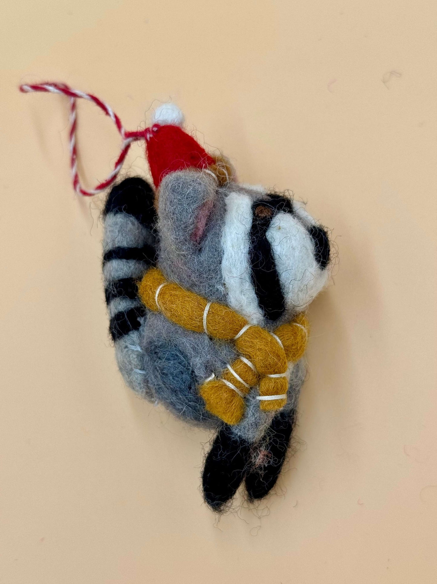 Handgemachter Filzanhänger in Form eines Weihnachtswaschbären mit Schal und Mütze, grau, schwarz und rot.